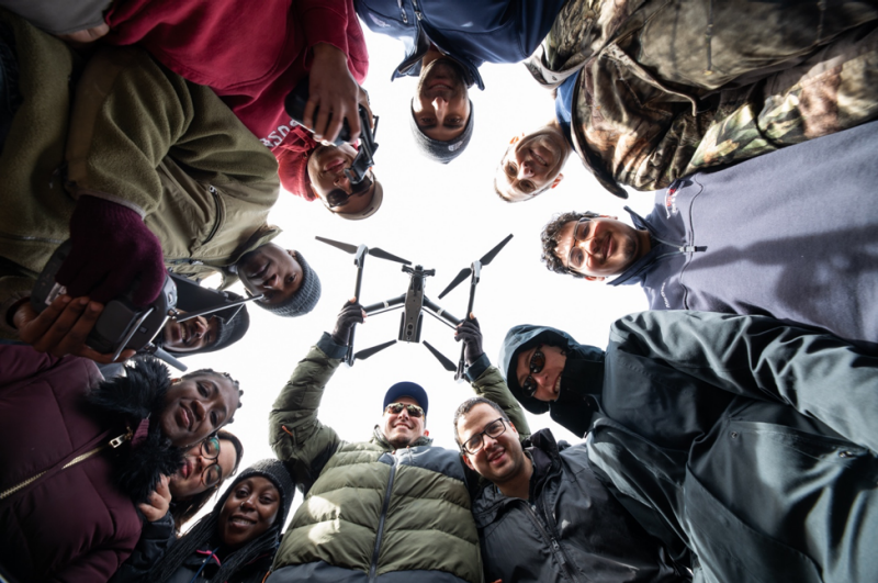 David Schmale (bottom, center) and Shane Ross (top, right) and their students are corralled in a circle and looking down at the camera. Schmale is holding a white unmanned aerial vehicle, also known as a drone. Peter Means for Virginia Tech.