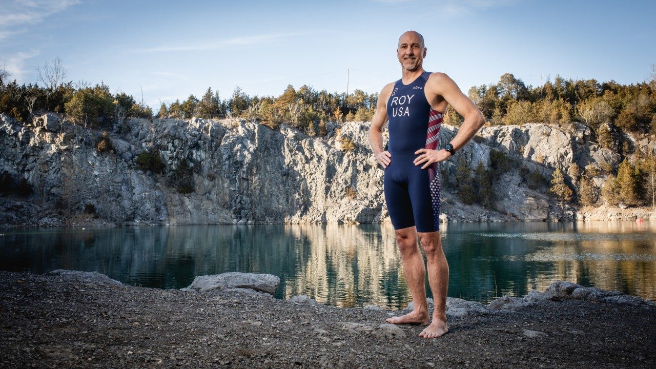 Chris Roy standing by a quarry filled with water in his USA triathlon wetsuit.